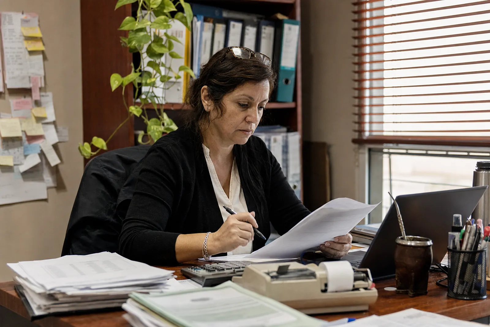 Persona trabajando con una laptop y papeles sobre el escritorio, con mate y termo al lado
